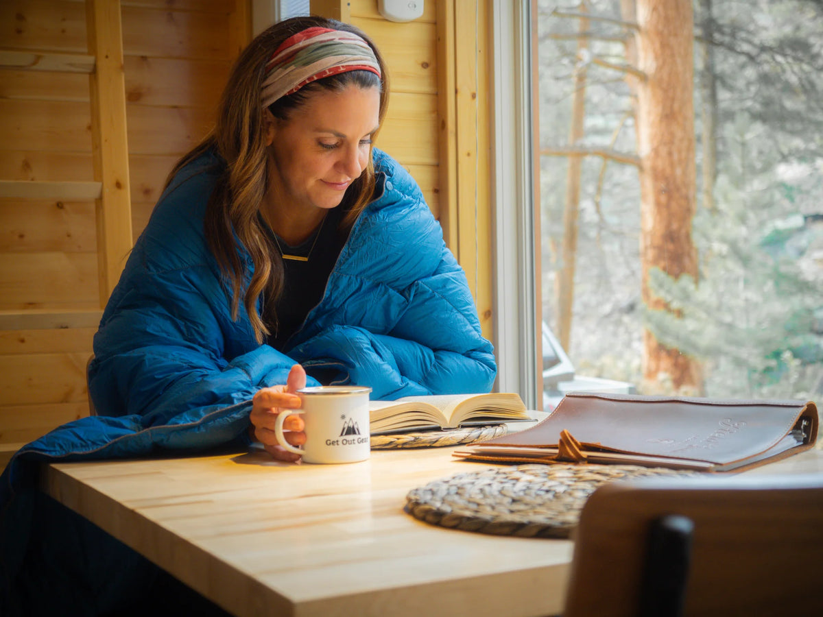 Woman in a blue jacket reading a book with a mug on a wooden table in a cozy cabin setting.