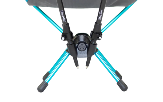 Close-up view of the base of a portable chair showing turquoise metal legs crossed and connected at a central black plastic hub, with two black chair poles labeled “Stay wild” and “Stay free.”.