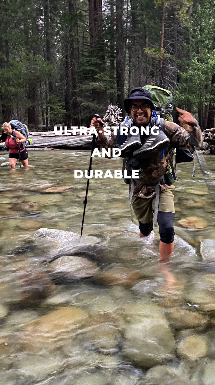 A smiling hiker with a backpack and trekking pole crosses a shallow, rocky river in the forest. The words ULTRA-STRONG AND DURABLE are overlaid across the center of the image. Another hiker is visible in the background.