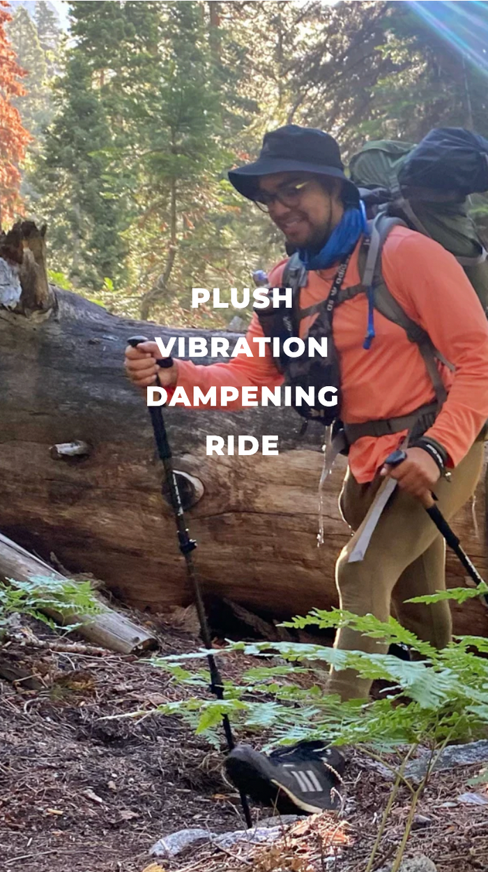 A man hiking in a forest with a hat, sunglasses, and backpack, using trekking poles. The text over the image reads: PLUSH VIBRATION DAMPENING RIDE. Sunlight filters through the trees.