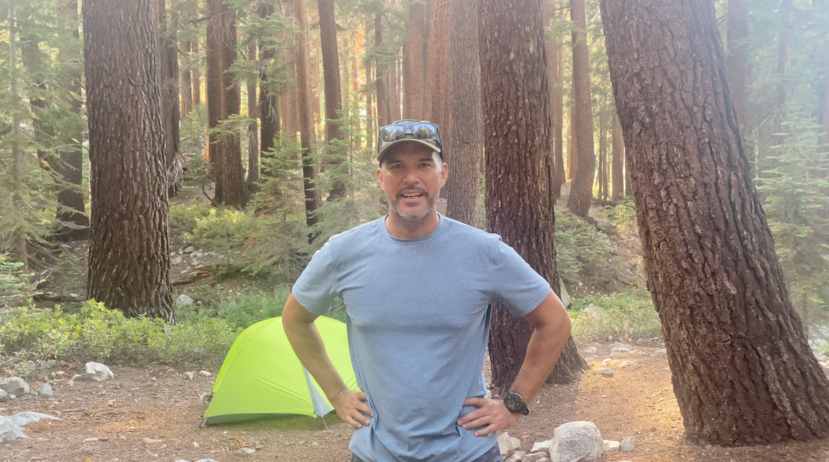 A man in a blue t-shirt stands with hands on hips in a forested campsite, smiling at the camera. Behind him is a green tent among tall pine trees, with sunlight filtering through the forest.