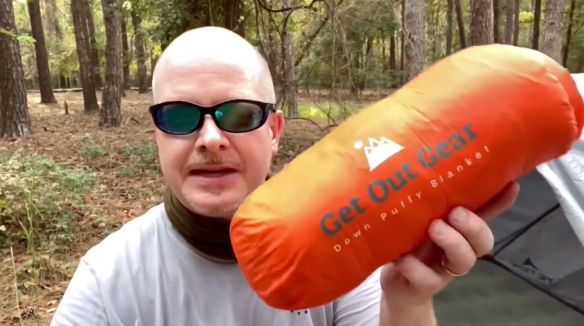 A person wearing sunglasses and a neck gaiter holds up an orange Get Out Gear Down Puffy Blanket bag in a forested outdoor setting.