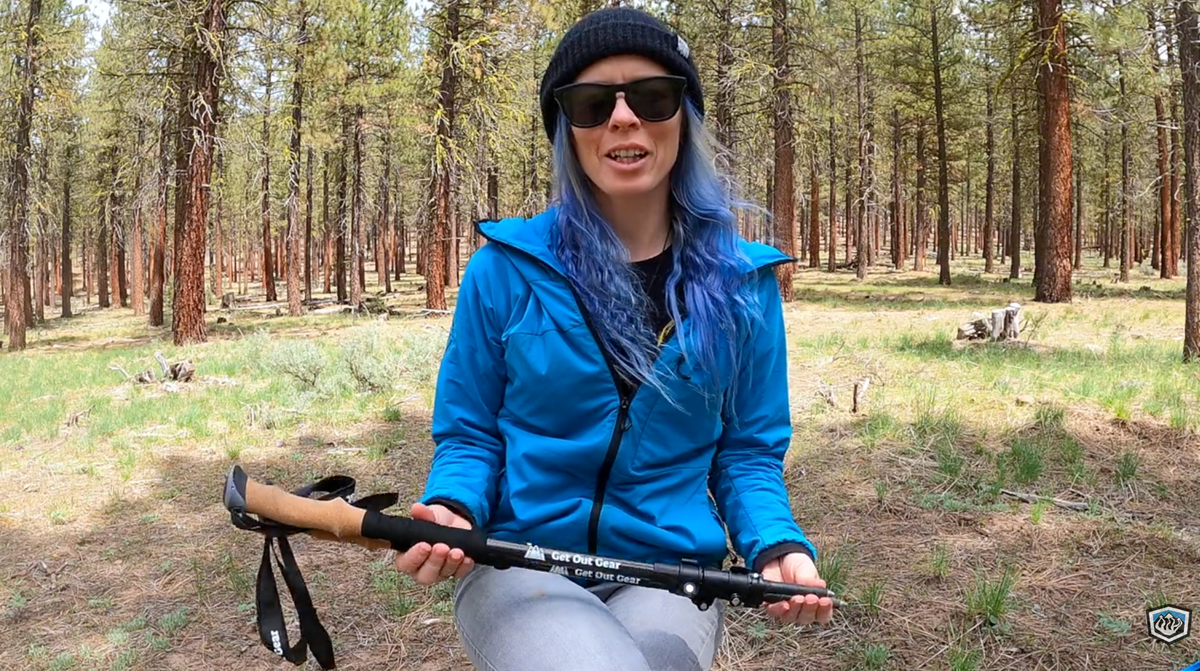 A person with long blue hair, wearing sunglasses, a black beanie, and a blue jacket, sits on the ground in a forest holding a pair of trekking poles. Pine trees surround them and sunlight filters through the trees.