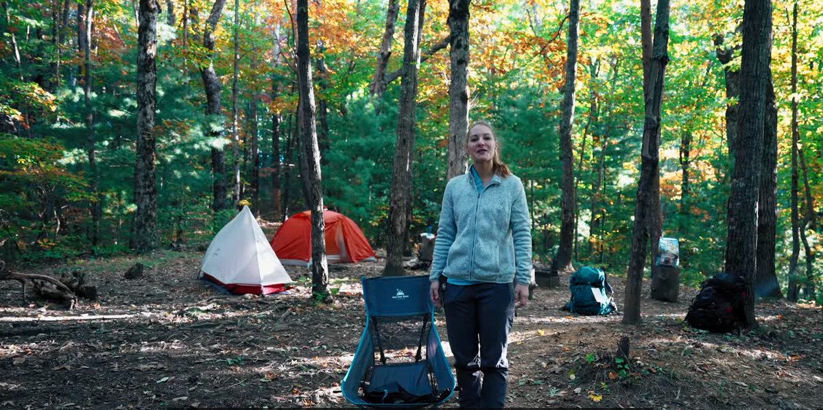 A woman stands in a wooded campsite with autumn foliage, two colorful tents, camping chairs, and backpacks visible among the trees. Sunlight filters through the leaves, creating a bright, peaceful outdoor scene.