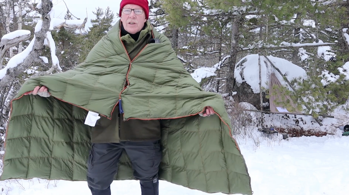 A person wearing glasses, a red hat, and winter clothes stands outdoors in the snow, holding open a large green blanket or quilt. Snow-covered trees and rocks are visible in the background.