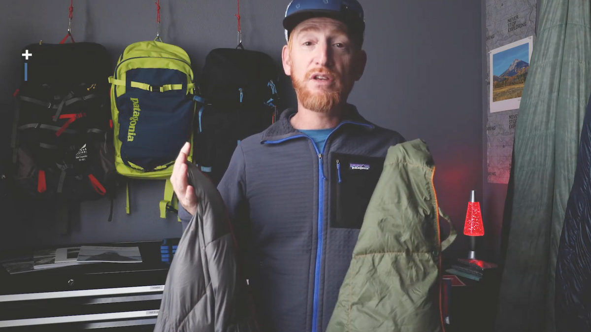 A man wearing a blue jacket and cap stands indoors holding two jackets, one gray and one green. Behind him are backpacks hanging on the wall, a red lava lamp, and outdoor-themed decor.