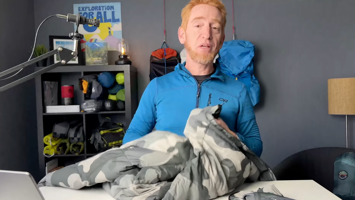 A man with reddish hair and a beard, wearing a blue zip-up jacket, holds a gray camouflage-patterned sleeping bag at a table in a room with outdoor gear on shelves and a blue backpack hanging on the wall behind him.