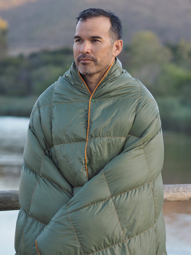 A man stands outdoors near a wooden railing, wrapped in a green quilted blanket. Trees, water, and hills are visible in the blurred background. He looks thoughtful and calm.